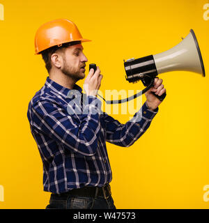 Angry worker man in orange helmet with a megaphone on yellow background - Image Stock Photo