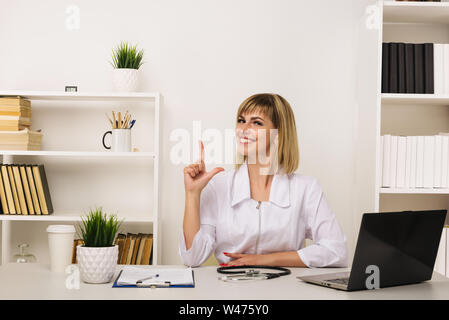 Friendly female doctor works at her desk in the office pointing up - image Stock Photo