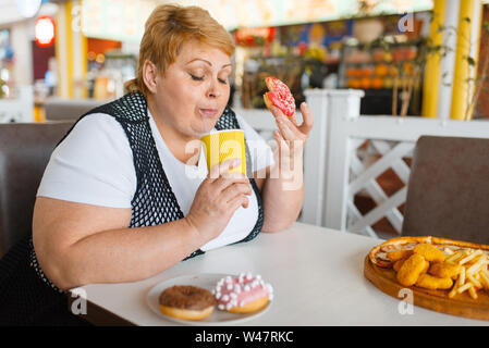 Fat woman eating doughnuts in fastfood restaurant, unhealthy food. Overweight female person at the table with junk dinner, obesity problem Stock Photo