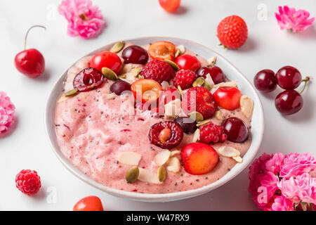 pink smoothie bowl or nice cream with fresh berries, rose flowers, nuts and seeds on white marble table. healthy breakfast. close up
