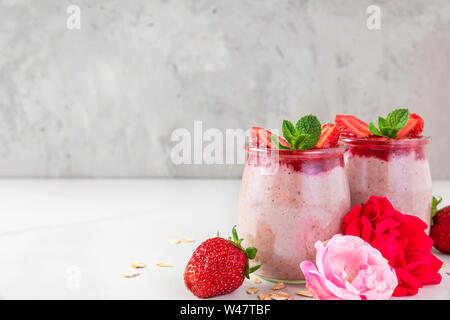 overnight oats or oatmeal porridge with fresh strawberries and mint in glasses with rose flowers on white marble table. healthy breakfast. close up Stock Photo