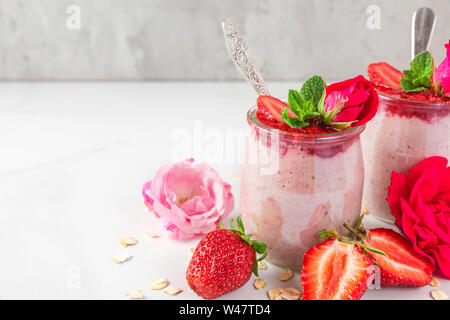 overnight oats or oatmeal porridge with fresh strawberries and mint in glasses with rose flowers and spoons on white marble table. healthy breakfast. Stock Photo