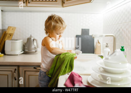 A little girl wipes the dishes on the kitchen. Baby doing housework at home. Young housewife clean the house Stock Photo