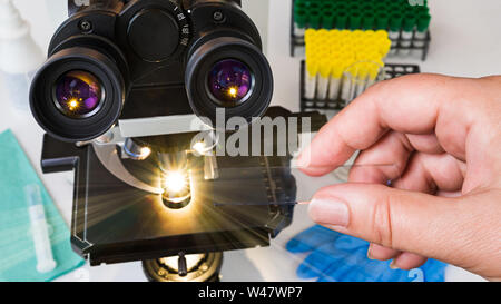 Laboratory optical microscope. Human hand holding a specimen on glass slide. Black medical scope, eyepieces, shining light rays and test tubes in rack. Stock Photo