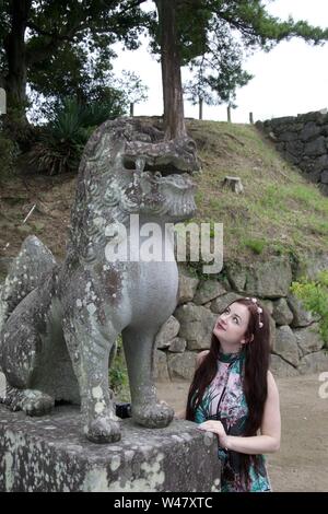 A fashionable, long haired, brunette, caucasian hippie girl looking up and admiring an ancient damaged Japanese statue of a mythical beast (dragon or Stock Photo