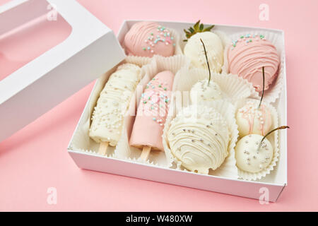 Strawberries, bananas, cherries and apricots coated with white and pink chocolate are lie in a gift box on a pink background Stock Photo