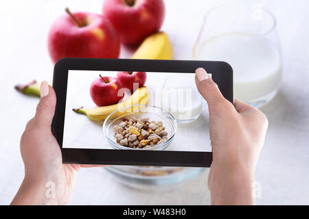 Female hands with a smartphone, on the screen of which is a photo of Healthy food with corn flakes cereal in a bowl, apple, banana and glass with milk Stock Photo