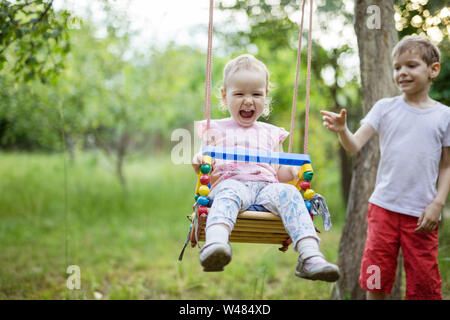 Young boy pushing toddler sister on swing. Siblings having fun outdoors on summer day.