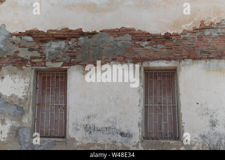 Worn brick wall of an old temple with vintage windows. Concrete wall with cracks showing red bricks. Stock Photo