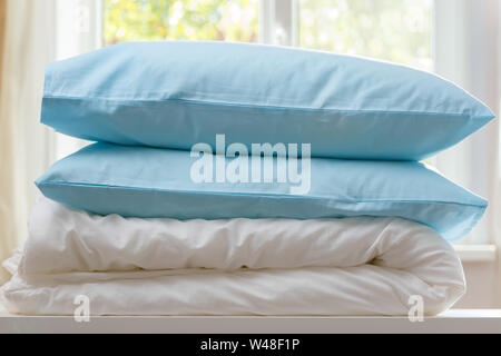 A folded duvet and pillows lie on a dresser against the background of a blurred window. Household. Stock Photo
