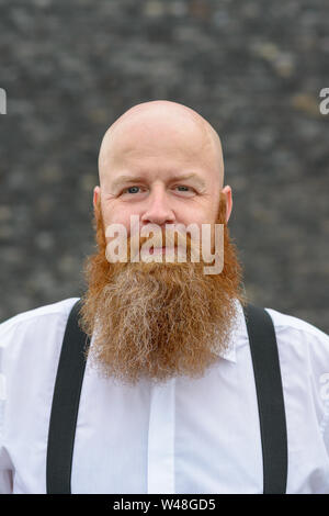 Portrait of a balding bearded redheaded man with a friendly smile wearing braces standing in front of a grey background Stock Photo