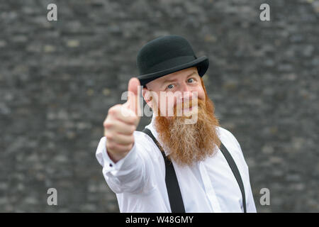 Motivated gleeful man with bushy red beard in retro bowler hat and braces giving a thumbs up gesture of success or support Stock Photo