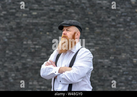 Overweight bearded man in cloth hat and braces standing with folded arms staring intently off to the left of the frame Stock Photo