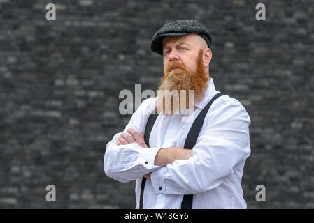 Overweight bearded man in cloth hat and braces standing with folded arms staring intently off to the left of the frame Stock Photo
