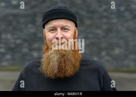Happy jovial bearded redhead man wearing a black beanie hat grinning at the camera with a mischievous expression Stock Photo