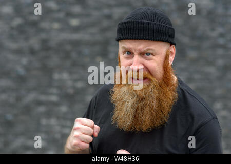 Angry aggressive man with bushy red beard in a knitted beanie hat making a fist as he snarls at the camera over grey Stock Photo