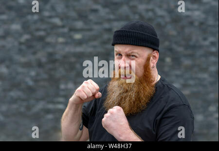 Angry aggressive man with bushy red beard in a knitted beanie hat making a fist as he snarls at the camera over grey Stock Photo