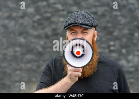 Bearded man wearing a cloth cap speaking into a megaphone making an announcement or public speaking Stock Photo
