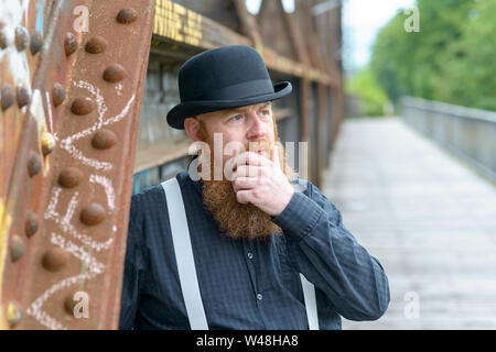 Thoughtful bearded man in a retro style bowler hat and braces standing leaning against an old bridge with his hand to his beard looking up Stock Photo