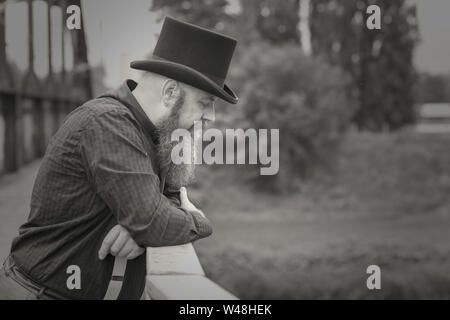 Black and White Photo of an bearded man in a top hat lost in deep thought as he stands leaning on the parapet of a bridge looking down into the river Stock Photo