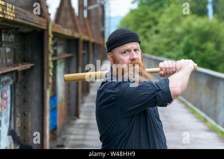 Threatening angry man with a red beard wearing a knitted beanie hat swinging a baseball bat over his shoulder while focusing his eyes on the camera Stock Photo