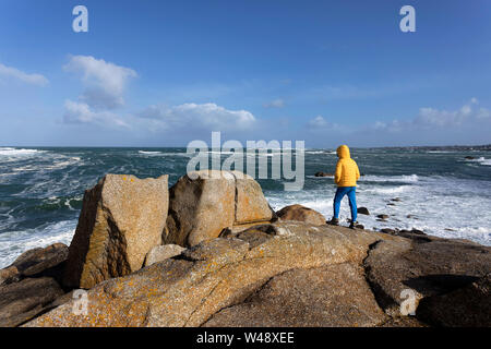 Young boy in yellow jacket standing on granite rocks watching big waves from the atlantic ocean on rugged coastal landscape in Brittany, France Stock Photo