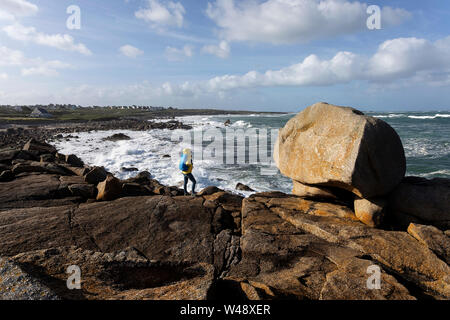 Woman in yellow jacket standing on granite rocks watching big waves from the atlantic ocean on rugged coastal landscape in Brittany, France Stock Photo