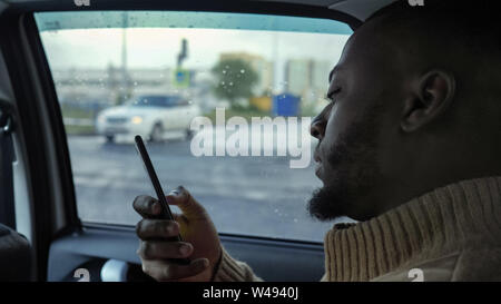 Portrait of black man is browsing smartphone. He is riding a car in rainy day in city. Afro american bearded guy is sitting in the back seat, side view. Stock Photo