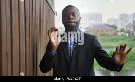 Afro american guy in earphones dances to favorite tune. Black man listens music in earbuds enjoys it in rainy street in city. He wears suit and shirt. Stock Photo
