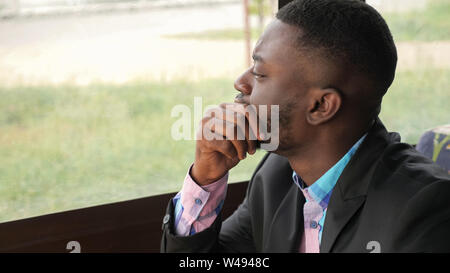 Afro american pensive tired young businessman is sitting in cafe. Portrait of young black man looks at window turns his head and looks at camera. Bearded young guy wears shirt and suit jacket. Stock Photo