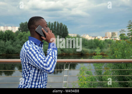 Afro american young guy is talking mobile phone, side view. Black man is calling on smartphone walking on river waterfront in city park in slow motion at overcast day. Stock Photo