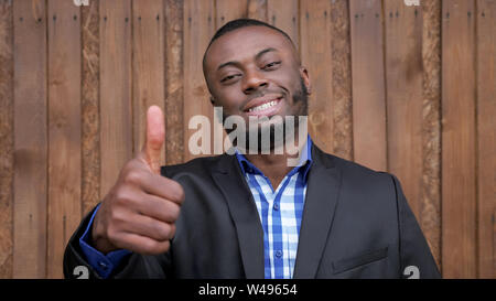 Afro american businessman in shirt and suit jacket, close-up. Black smiling man shows gesture thumb up on dark wood background. Winner concept. Stock Photo