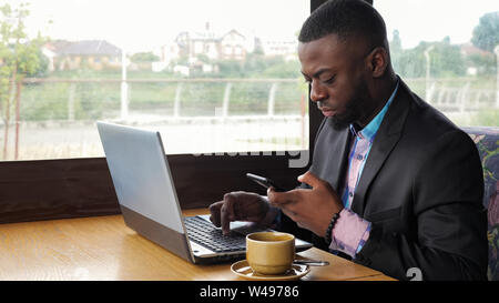 Afro american man has breakfast. Black businessman works on laptop with smartphone in cafe and drinks coffee. He types on computer looking at mobile phone screen. Wears in shirt and suit jacket. Stock Photo