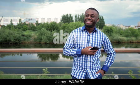 Young man with phone smiling. Afro american guy stands near fence on river waterfront in city park. He wears plaid shirt and jeans. Stock Photo