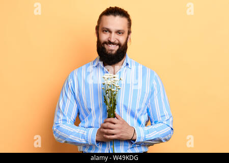 smiling kind bearded stylish man holding field flowers in his hands and looking at the camera. close up portrait. surprise, man going to give a presen Stock Photo