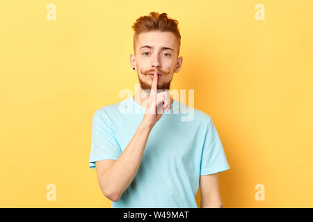 handsome calm stylish man wearing casual fashion t-shirt holding index finger at his lips, showing shh gesture, asking to keep silent and not to make Stock Photo