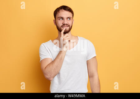 pensive cunning young man touching chin with finger thinking or considering. making a plan, looking up. close up portrait. headshot, isolated yellow b Stock Photo