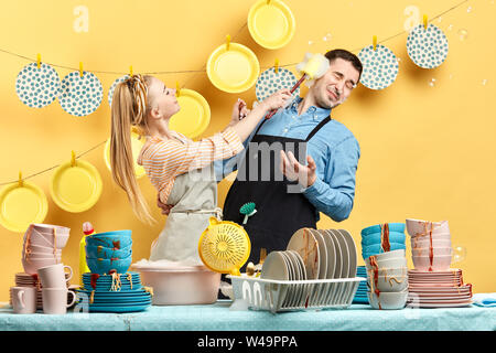 childish pleasant cheerful girl smearing her boyrirnd's face with foam, they are standing behind table with dirty plates, bowls and cups, close up pho Stock Photo