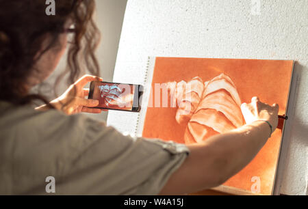Woman using mobile phone photo reference to drawing a picture at home in front of a bright window. Using the technology to paint. Stock Photo
