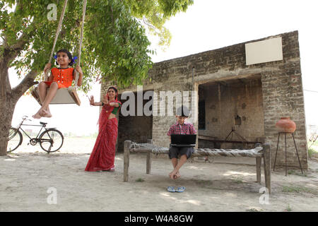 Indian woman pushing her daughter on a rope swing with her son using laptop Stock Photo