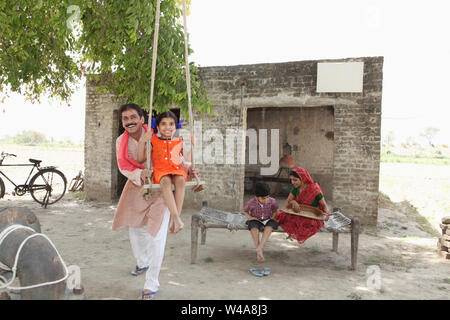 Man pushing his daughter on the swing with his wife cleaning grain Stock Photo