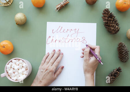 Female hands signing Christmas card with pen at green table with Christmas decorations and treats Stock Photo