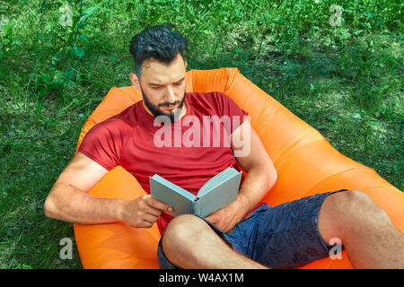 Young male student, resting on the nature with a book in his hands, lying in an inflatable lounger or air sofa. Stock Photo