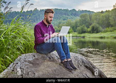 A young bearded man sits on a stone near the river with a laptop in his lap, he looks at the screen and types text on the keyboard. Stock Photo