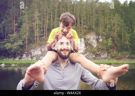 The family walks through the forest near the river, the father carries his son on his shoulders, eco-tourism, outdoor recreation during the summer hol Stock Photo