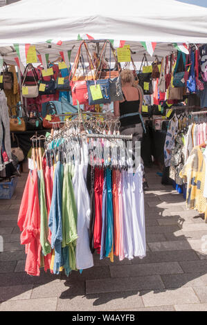 Brightly coloured colthes on racks at front of weekly market stall and lady looking at bags at the back of the stall Stock Photo