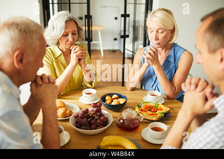 Group of senior people praying while sitting around table in kitchen, copy space Stock Photo