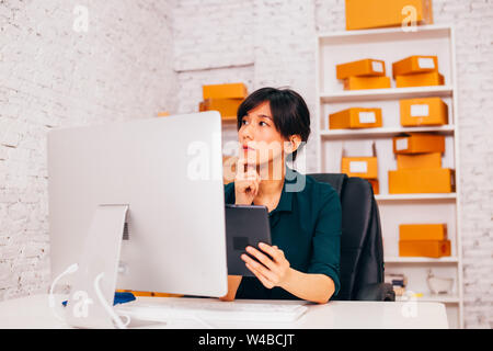 Asian entrepreneur with tablet looking away and thinking while sitting at desk in office of e commerce company Stock Photo
