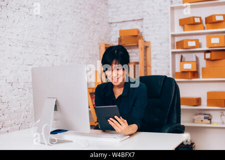 Cheerful Asian businesswoman smiling and browsing tablet while sitting at desk with computer in office Stock Photo