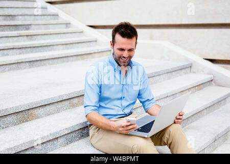 Handsome student sitting on stone steps and using a laptop and mobile Stock Photo